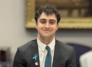 Photo of Ian Michelson.  Ian is wearing a blazer, white collared shirt, teal tie, and teal sexual assault awareness ribbon.